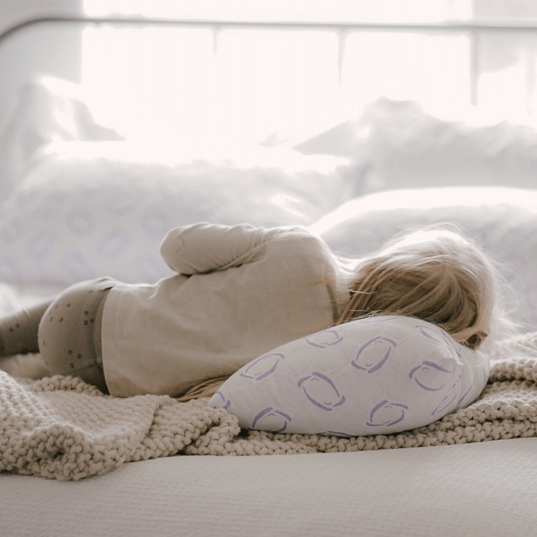 Child lying on a bed with a white pillow featuring purple patterns

